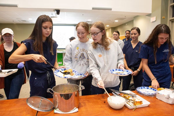 A white-haired woman pours food from a pot onto a paper plate as two women look on and others stand in line behind them.