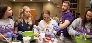 A bearded man in a purple T-shirt talks to a group of women preparing food in a large meeting room.