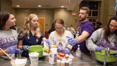 A bearded man in a purple T-shirt talks to a group of women preparing food in a large meeting room.