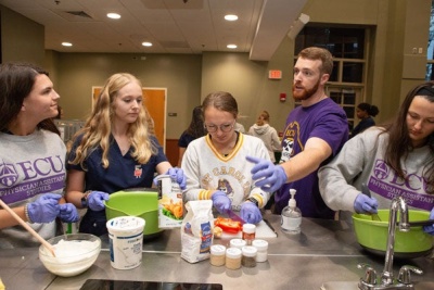 A bearded man in a purple T-shirt talks to a group of women preparing food in a large meeting room.